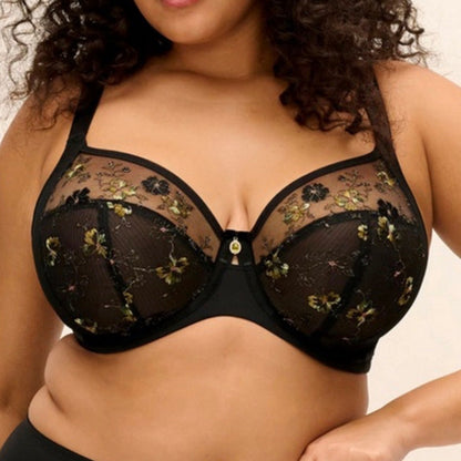 A plus-size woman with curly dark hair wearing a black sheer floral lace bra, photographed against a plain light beige background.