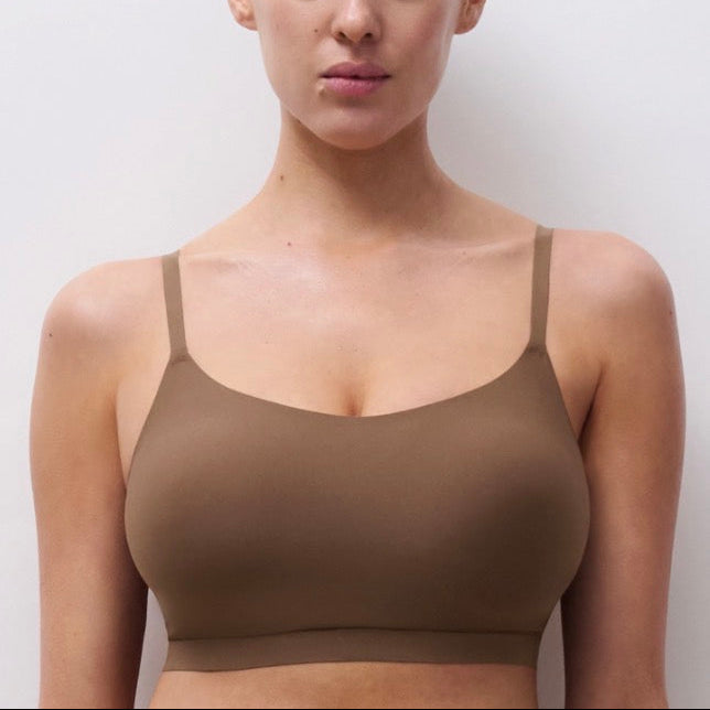 A woman wearing a seamless, solid brown bralette with thin straps, shown from the chest up against a plain light gray background.