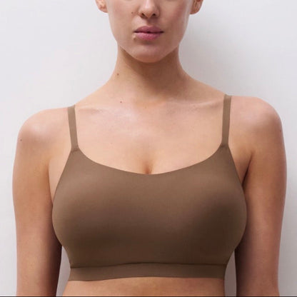 A woman wearing a seamless, solid brown bralette with thin straps, shown from the chest up against a plain light gray background.