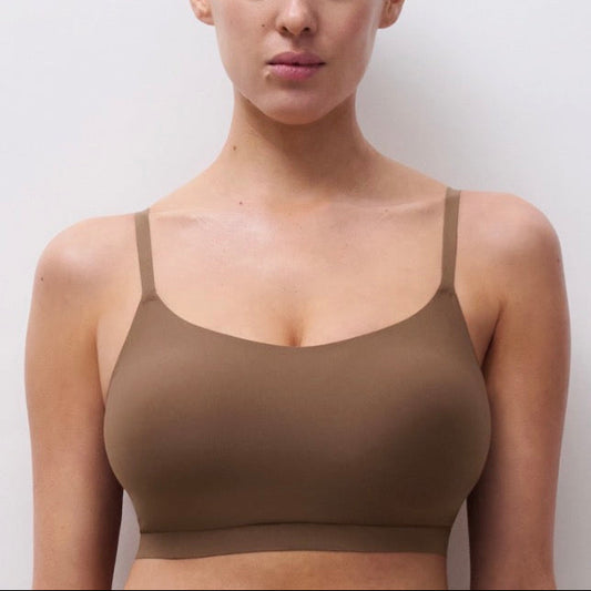 A woman wearing a seamless, solid brown bralette with thin straps, shown from the chest up against a plain light gray background.