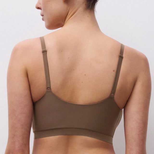 A woman shown from the back wearing a solid taupe-brown bralette with thin adjustable shoulder straps, set against a plain light gray studio background.