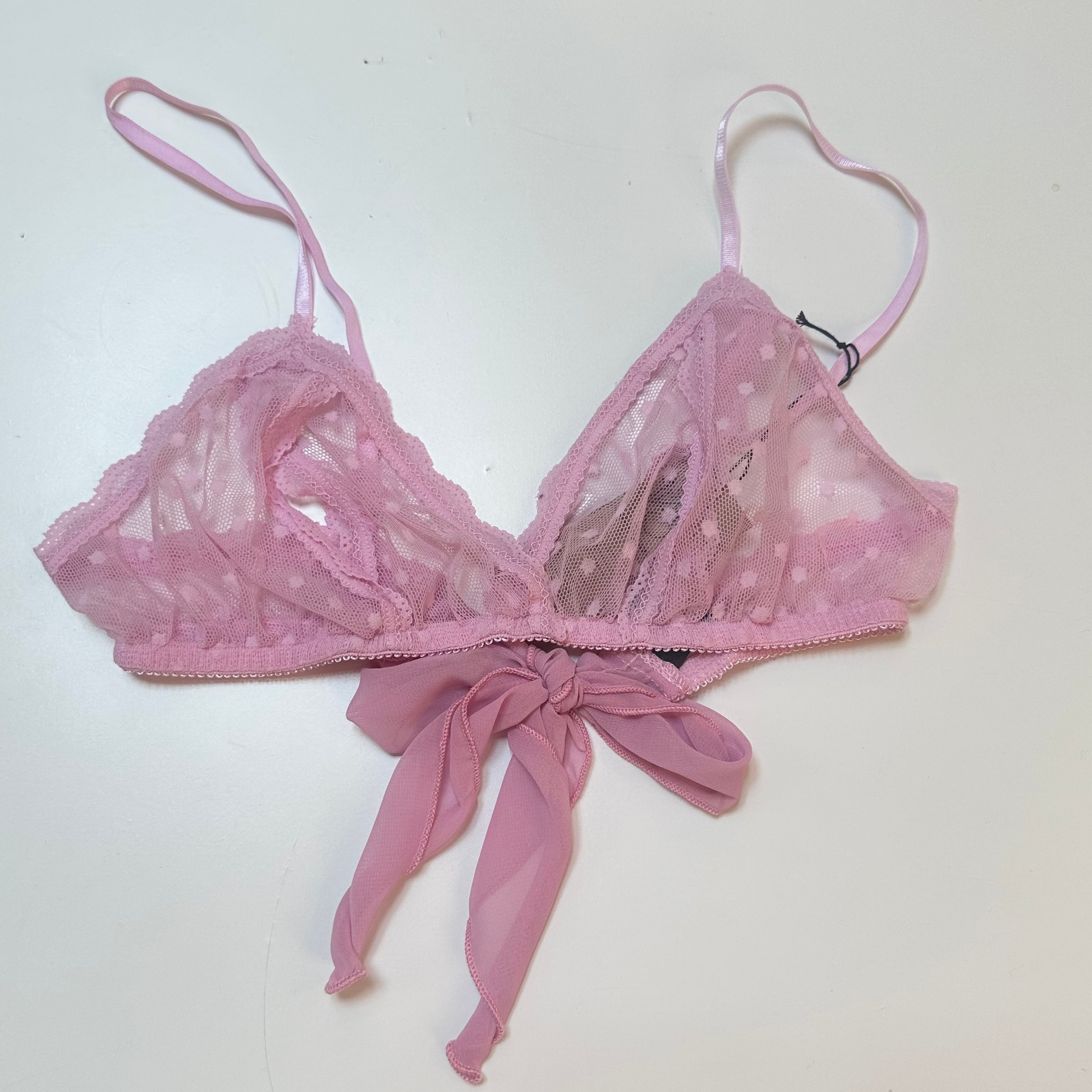 A pink sheer mesh bralette with lace cups, thin adjustable straps, and a matching fabric bow at the center front, laid flat on a white surface.