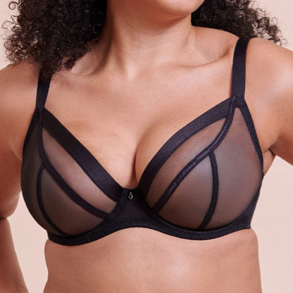 A smiling woman with curly dark hair wears a black sheer mesh and structured bra against a plain beige studio background.