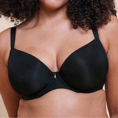 A woman with voluminous curly black hair wearing a black wireless underwire bra against a solid beige background.