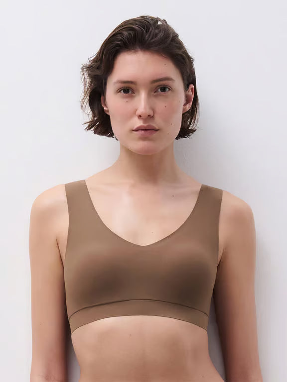 A woman with short brown hair wearing a seamless, scoop-neck bralette in a solid taupe-brown color, photographed against a clean, light gray studio background.